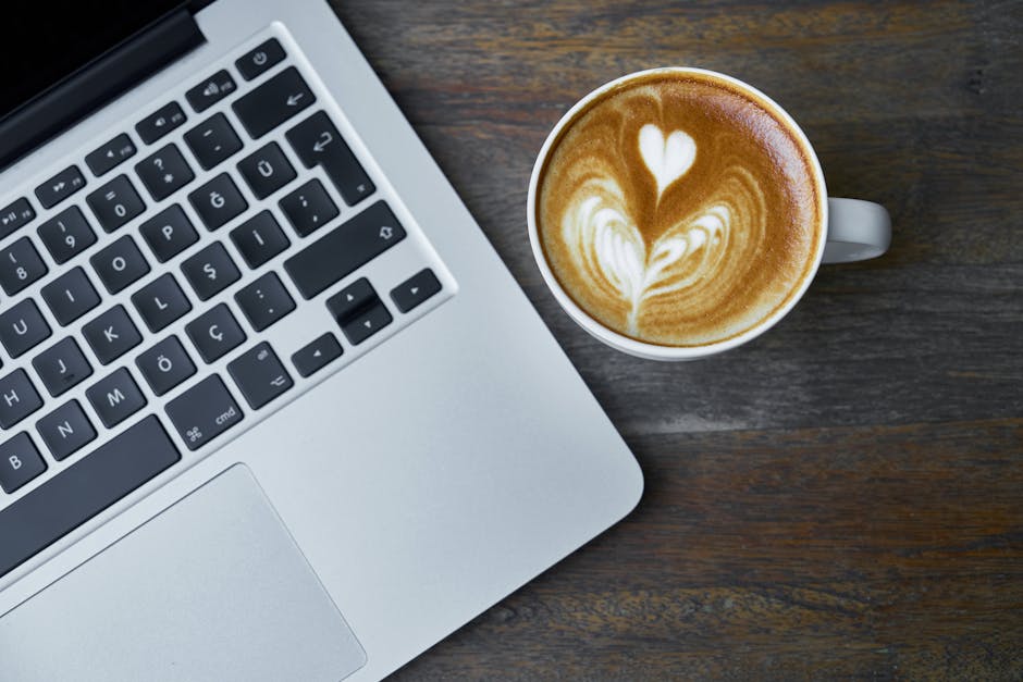 A overhead view of a cappuccino with heart-shaped latte art next to a laptop on a wooden table.